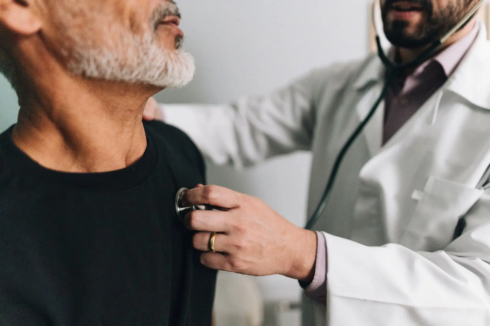 A physician in a white coat listens to an older man’s heart with a stethoscope during an office exam.