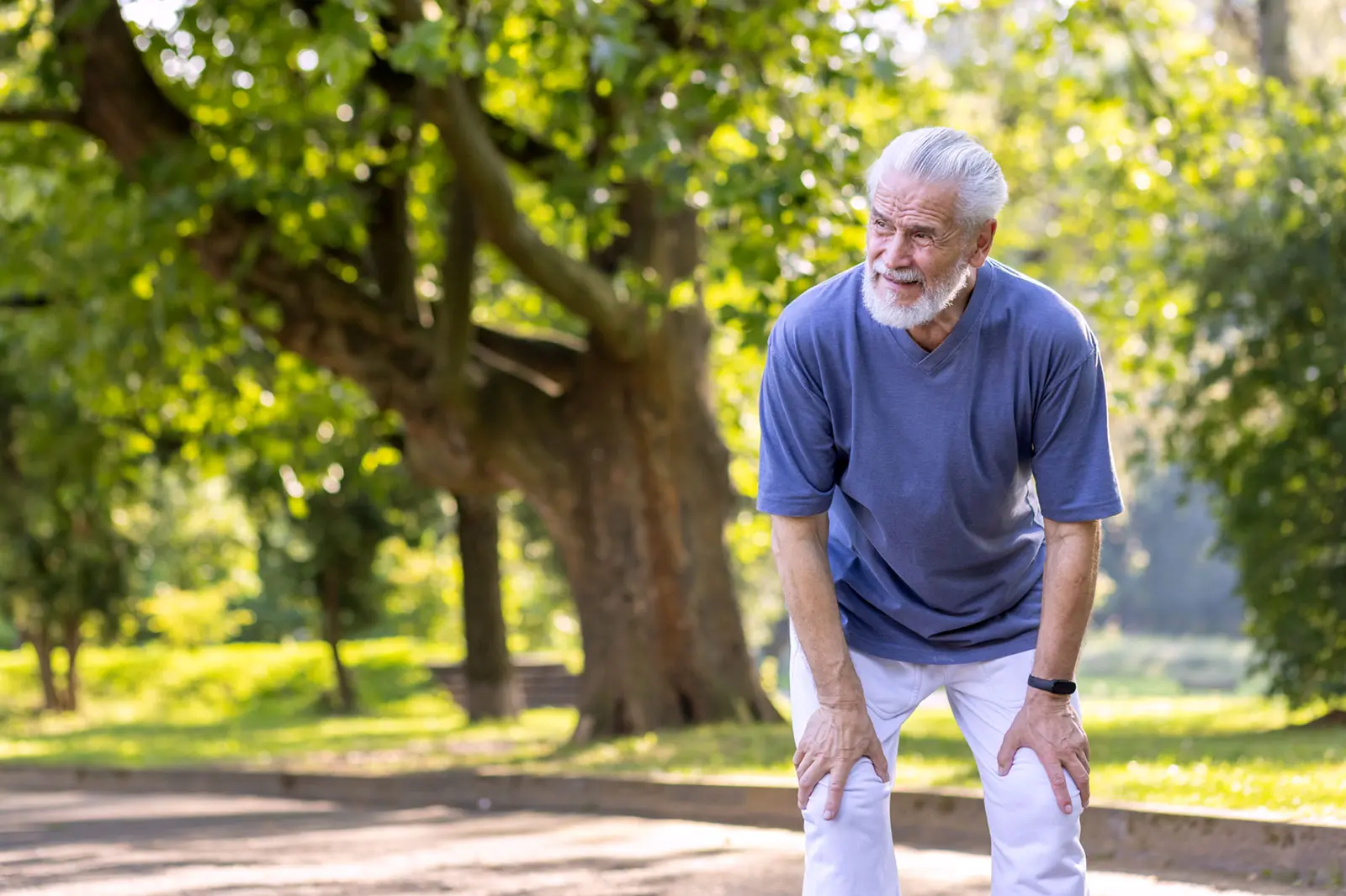 An older man in a park stops during a walk and rests his hands on his knees to catch his breath.